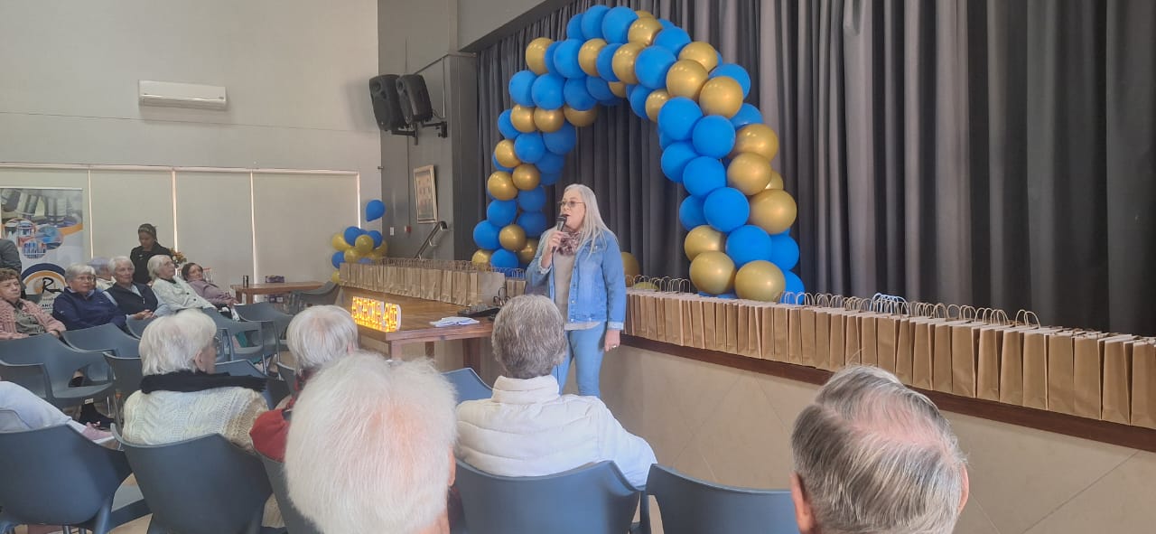 Karen addressing a seated audience beneath a blue and gold balloon arch